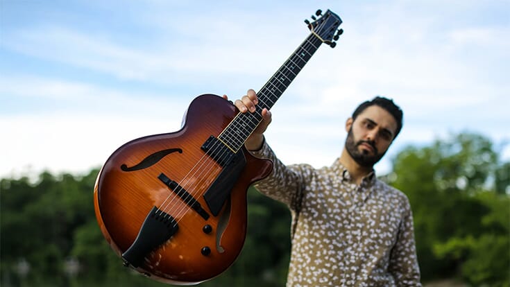 Adam Moezinia outdoors, holding up a guitar with one hand