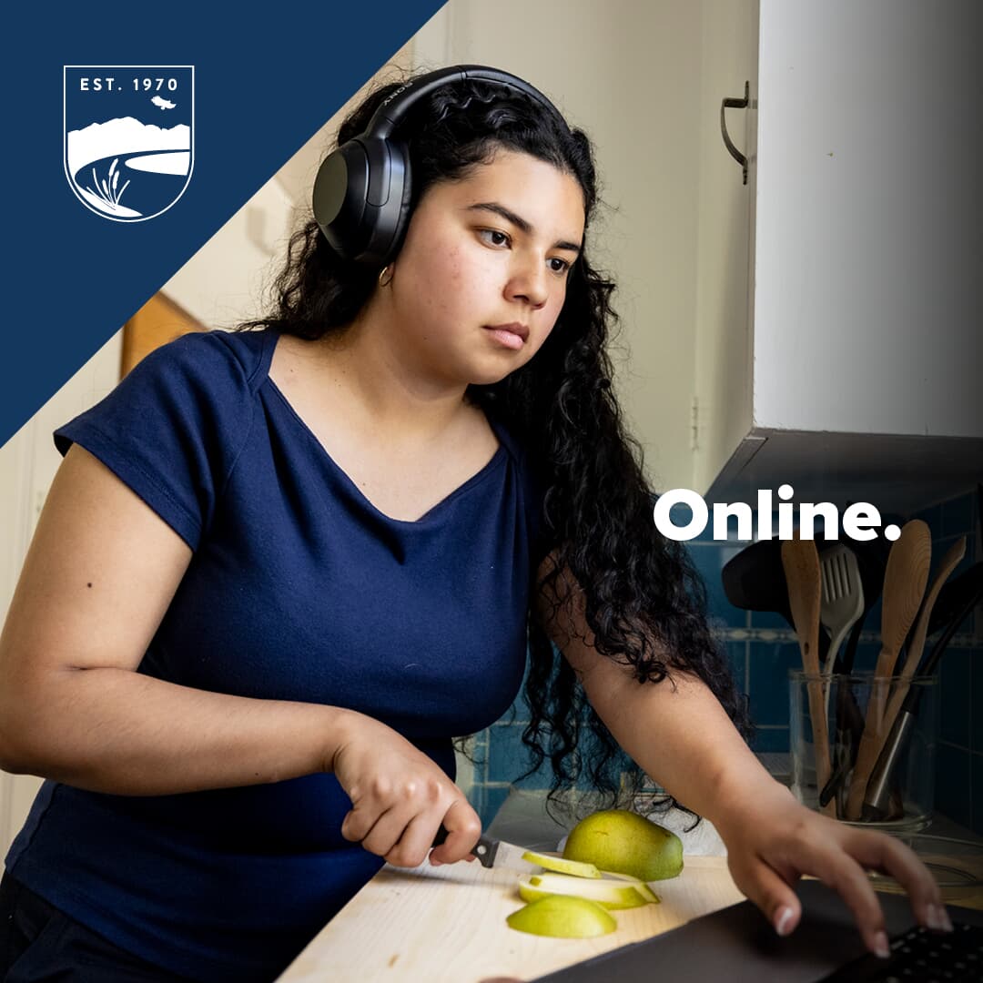 Text that reads Online alongside Cosumnes River College logo and photo of young woman cutting an apple in her kitchen while looking at a laptop